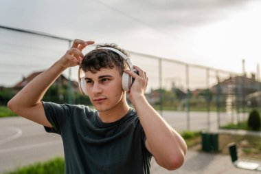 Beyaz tenli genç bir erkek açık hava eğitim parkı spor salonunda duruyor. Kulaklıklı cep telefonu kullanıyor. Gerçek insanları eğitmek için internetten mesajlar yolluyor.