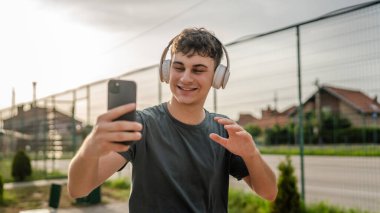 Beyaz tenli genç bir erkek açık hava eğitim parkı spor salonunda duruyor. Kulaklıklı cep telefonu kullanıyor. Gerçek insanları eğitmek için internetten mesajlar yolluyor.