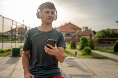 Beyaz tenli genç bir erkek açık hava eğitim parkı spor salonunda duruyor. Kulaklıklı cep telefonu kullanıyor. Gerçek insanları eğitmek için internetten mesajlar yolluyor.