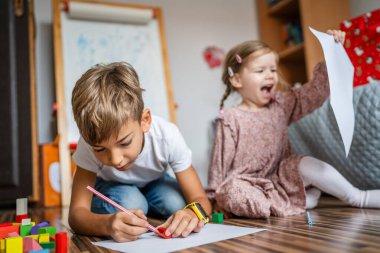 Küçük beyaz bir oğlan ve kız kardeşi evde pastel boyayla resim çiziyor.