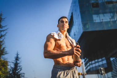 Beyaz tenli genç bir erkek sporcu açık hava spor salonunda çalışırken fren yapıyor. Elinde destek malzemesi tutuyor. Kendine güveni tam, güçlü, güçlü bir kopya alanı var.