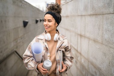Genç bir kadın yetişkin kız öğrenci sınıfa gider dizüstü bilgisayarlı bir fincan kahve ve iş malzemesi taşır. Gerçek insan utangaç, mutlu, kulaklıklı, kendine güvenen bir gülümseme.
