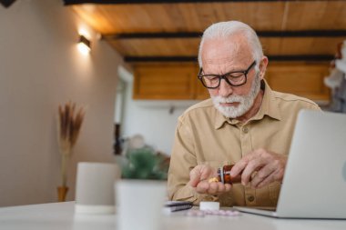 Son sınıf beyaz erkek büyükbaba evinde oturup ağrı kesici ya da vitamin takviyesi paketini tutuyor. İlaç almaya hazır.