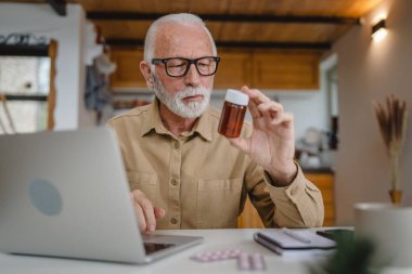 Son sınıf beyaz erkek büyükbaba evde oturup ağrı kesici veya vitamin takviyesi paketlerini tutuyor. Bilgisayardan sipariş edilen dizüstü bilgisayarın yanında fotokopi için hazır bekliyor.