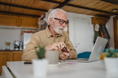 Son sınıf beyaz erkek büyükbaba evde oturup ağrı kesici veya vitamin takviyesi paketlerini tutuyor. Bilgisayardan sipariş edilen dizüstü bilgisayarın yanında fotokopi için hazır bekliyor.