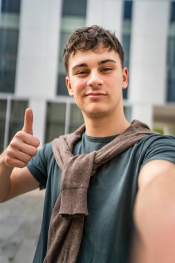 Genç, yetişkin, beyaz bir genç, dışarıda durup fotoğraf çekiyor. Kameraya bakıyor. Kendine güveni tamdır.