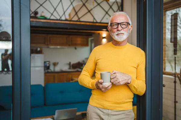 One man senior caucasian male with beard and eyeglasses stand on balcony at door in day happy smile hold cup of coffee copy space morning daily routine concept