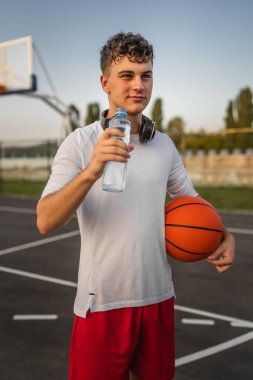 Kafkasyalı ergen, açık havada güneşli bir günde plastik bir şişe su açarken delik basketbolu oynuyor.