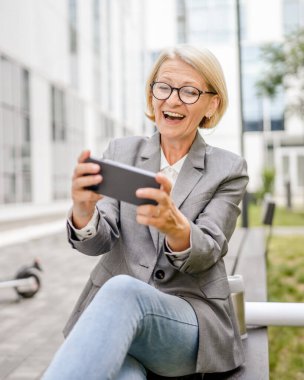 Olgun, sarışın, beyaz bir kadın açık havada mutlu bir şekilde oturup video oyunları oynuyor, cep telefonu kullanıyor, eğleniyor akıllı telefon kullanıyor, internet üzerinden fotokopi çekiyor, gözlük takıyor ve tişört giyiyor.