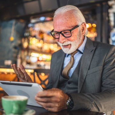 Bir üst düzey Kafkas erkek iş adamı dijital tablet kullanıyor kafede oturup görüntülü konuşma bağlantısı kuruyor ve iletişim kıyafeti fotokopi odası giyiyor.