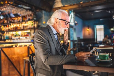 Bir üst düzey Kafkas erkek iş adamı dijital tablet kullanıyor kafede oturup görüntülü konuşma bağlantısı kuruyor ve iletişim kıyafeti fotokopi odası giyiyor.
