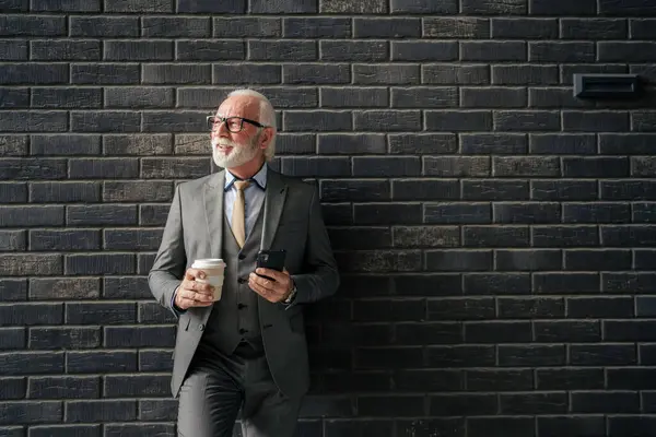one senior man businessman wear suit with gray beard stand outdoor in front of brick wall of the company hold smartphone and coffee entrepreneur or executive copy space