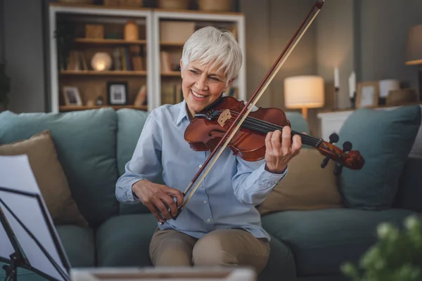Mature senior caucasian woman learn to play violin practice at home online professor teacher teach or have internet private class on digital tablet while sit on sofa bed at home active senior hobby