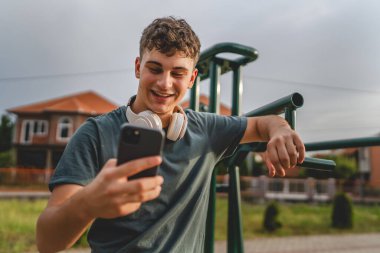 Beyaz tenli genç bir erkek açık hava eğitim parkı spor salonunda duruyor. Kulaklıklı cep telefonu kullanıyor. Gerçek insanları eğitmek için internetten mesajlar yolluyor.