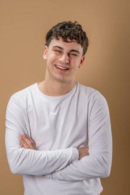 20 yaşında beyaz bir adamın portresi. Badem rengi stüdyo arkaplanının önünde kameraya bakıyor. Gülümsüyor.