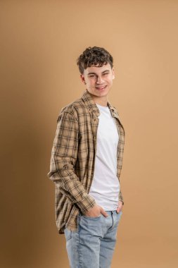 20 yaşında beyaz bir adamın portresi. Badem rengi stüdyo arkaplanının önünde kameraya bakıyor. Gülümsüyor.