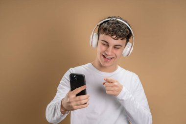 Beyaz tenli genç bir adam bej arka planda çekim yapıyor. Kulaklıklı cep telefonu kullanıyor. Müzik mesajlar gönderiyor ya da internetteki uygulamaları kopyalıyor.