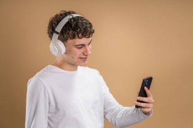 Beyaz tenli genç bir adam bej arka planda çekim yapıyor. Kulaklıklı cep telefonu kullanıyor. Müzik mesajlar gönderiyor ya da internetteki uygulamaları kopyalıyor.