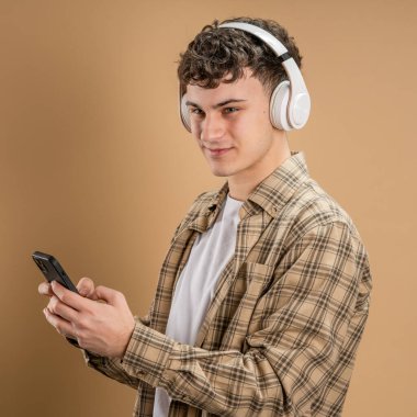 Beyaz tenli genç bir adam bej arka planda çekim yapıyor. Kulaklıklı cep telefonu kullanıyor. Müzik mesajlar gönderiyor ya da internetteki uygulamaları kopyalıyor.