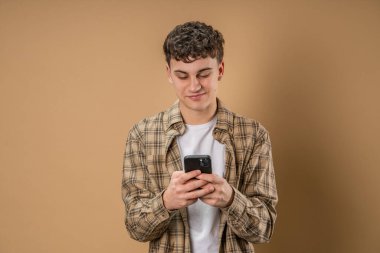 Beyaz tenli bir erkek çocuk internetteki sosyal ağları taramak ya da SMS mesajları atmak için akıllı cep telefonu kullanıyor. Stüdyo resmi, bej arka plan gülüşü.