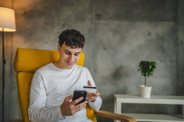 Genç bir beyaz erkek evde oturuyor. İnternetten kredi kartı ve dizüstü bilgisayar alışverişi yapıyor. İnternet üzerinden modern e-bankacılık cep telefonu ve kredi kartı satın alıyor.