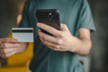 Genç bir beyaz erkek evde oturuyor. İnternetten kredi kartı ve dizüstü bilgisayar alışverişi yapıyor. İnternet üzerinden modern e-bankacılık cep telefonu ve kredi kartı satın alıyor.