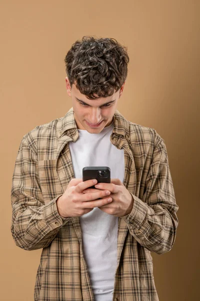 Beyaz tenli bir erkek çocuk internetteki sosyal ağları taramak ya da SMS mesajları atmak için akıllı cep telefonu kullanıyor. Stüdyo resmi, bej arka plan gülüşü.