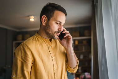 Yetişkin bir beyaz erkek cep telefonu akıllı telefonu kullanarak evde konuşma yapıyor. Tişört ve gözlük takıyor. Gerçek insan mekanı kopyalıyor.