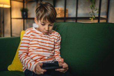 Bir çocuk evde oturuyor cep telefonunu tutuyor akıllı telefon izliyor.