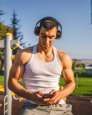 Beyaz tenli genç bir erkek açık hava eğitim parkı spor salonunda duruyor. Kulaklıklı cep telefonu kullanıyor. Gerçek insanları eğitmek için internetten mesajlar yolluyor.