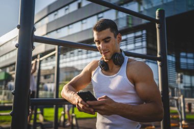 Beyaz tenli genç bir erkek açık hava eğitim parkı spor salonunda duruyor. Kulaklıklı cep telefonu kullanıyor. Gerçek insanları eğitmek için internetten mesajlar yolluyor.