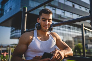 Beyaz tenli genç bir erkek açık hava eğitim parkı spor salonunda duruyor. Kulaklıklı cep telefonu kullanıyor. Gerçek insanları eğitmek için internetten mesajlar yolluyor.