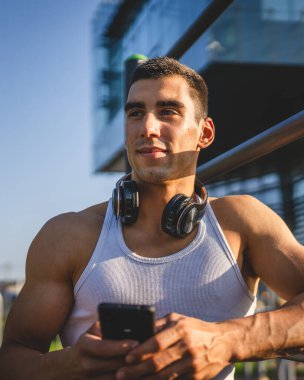 Beyaz tenli genç bir erkek açık hava eğitim parkı spor salonunda duruyor. Kulaklıklı cep telefonu kullanıyor. Gerçek insanları eğitmek için internetten mesajlar yolluyor.