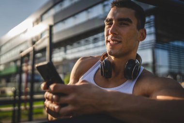 Beyaz tenli genç bir erkek açık hava eğitim parkı spor salonunda duruyor. Kulaklıklı cep telefonu kullanıyor. Gerçek insanları eğitmek için internetten mesajlar yolluyor.