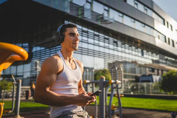 One man caucasian young male stand at outdoor open training park gym use mobile phone smartphone with headphones send messages texting or browse internet online app for training real person copy space