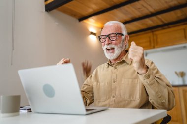 Sakallı ve gözlüklü yaşlı bir beyaz adam dizüstü bilgisayarda çalışıyor. Evde mutlu bir gülümseme başarısı var.
