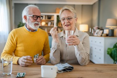 Üst düzey çift kadın ilaç ya da vitamin tedavisi alırken kocası beyaz adam evde masada oturuyor. Gerçek insanlar, aile emekliliği ve sağlık konsepti.