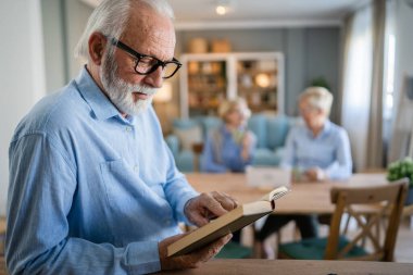 Yetişkin, yaşlı, beyaz, emekli, modern, kısa saçlı ve sakallı bir adam evde kitap okuyor. Gerçek gözlük takıyor.