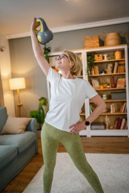 Evde kettlebell Girya Rus çanıyla antrenman yapan olgun bir beyaz kadın. Dairesinde sağlıklı yaşam tarzı kavramında kadın egzersizi yapıyor.