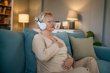 Yetişkin bir yaşlı beyaz kadın internet rehberli meditasyon için kulaklık kullanıyor. Farkındalık yogası yapıyor. Evde gözleri kapalı.