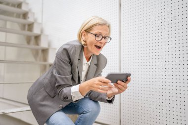 Olgun, sarışın, beyaz bir kadın açık havada oturup mutlu bir gülümseme alıyor video oyunları oynuyor, eğleniyor cep telefonu kullanıyor, akıllı telefon kullanıyor, internet üzerinden gözlük takıyor.