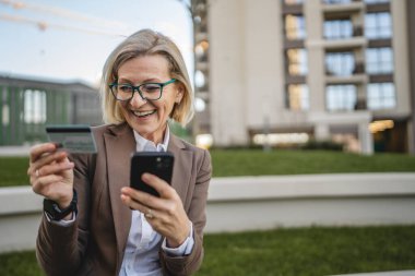 Bir kadın binanın önünde oturmuş cep telefonuna bakıyor, elinde kredi kartı tutuyor ve internetten ödeme yapıyor.