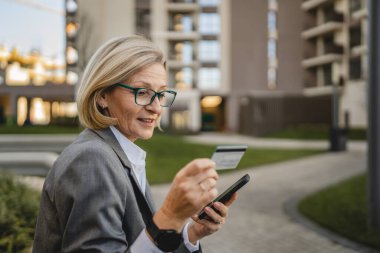 Bir kadın binanın önünde oturmuş cep telefonuna bakıyor, elinde kredi kartı tutuyor ve internetten ödeme yapıyor.