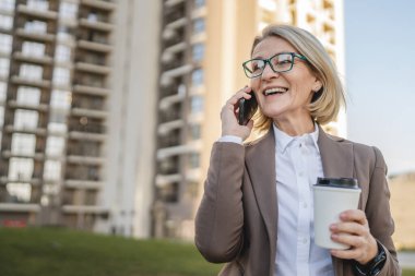 Olgun bir kadın binaların önünde cep telefonuyla konuşuyor ve elinde kahve, modern iş ve yaşam tarzı konsepti tutuyor.