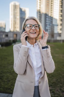 Binaların önünde cep telefonuyla konuşan olgun bir kadın, modern iş ve yaşam tarzı kavramı.