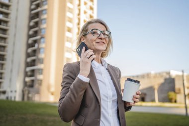 Olgun bir kadın binaların önünde cep telefonuyla konuşuyor ve elinde kahve, modern iş ve yaşam tarzı konsepti tutuyor.