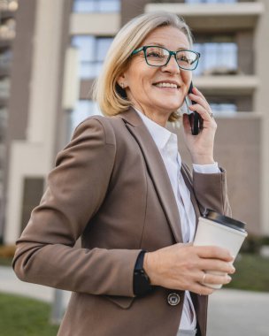 Olgun bir kadın binaların önünde cep telefonuyla konuşuyor ve elinde kahve, modern iş ve yaşam tarzı konsepti tutuyor.