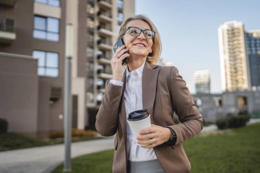 Olgun bir kadın binaların önünde cep telefonuyla konuşuyor ve elinde kahve, modern iş ve yaşam tarzı konsepti tutuyor.