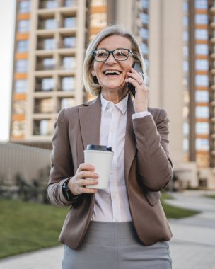 Olgun bir kadın binaların önünde cep telefonuyla konuşuyor ve elinde kahve, modern iş ve yaşam tarzı konsepti tutuyor.