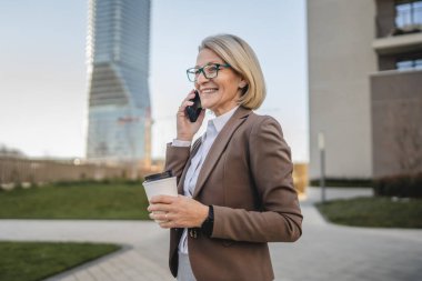 Olgun bir kadın binaların önünde cep telefonuyla konuşuyor ve elinde kahve, modern iş ve yaşam tarzı konsepti tutuyor.